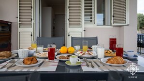 Ett frukostbord dukat med olika rätter, inklusive bakverk, frukter och drycker, på en balkong med öppna fönsterluckor och utsikt över interiören.