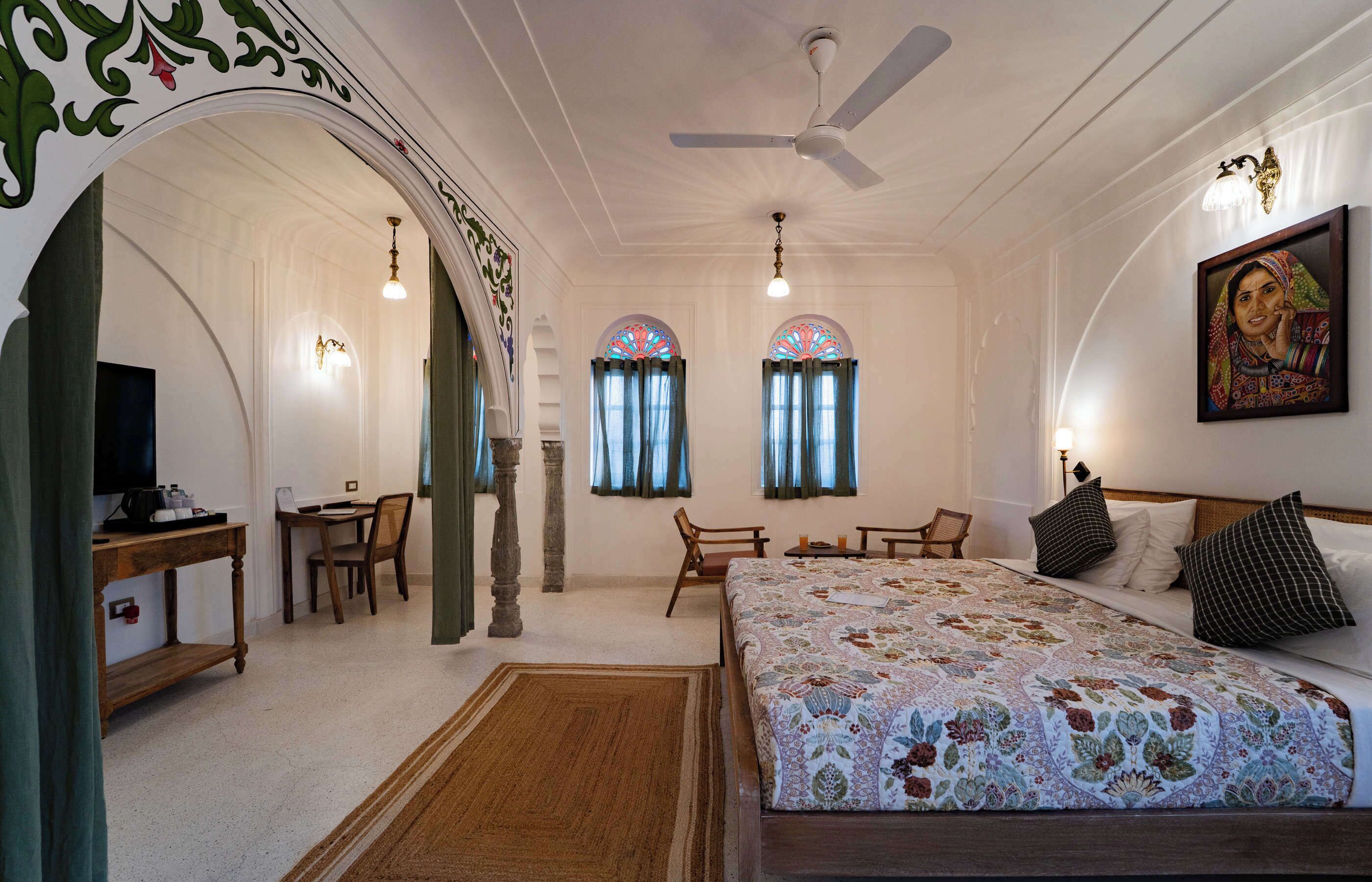 Una habitación de hotel con una cama grande, un escritorio, dos sillas, un ventilador de techo y un cuadro en la pared.