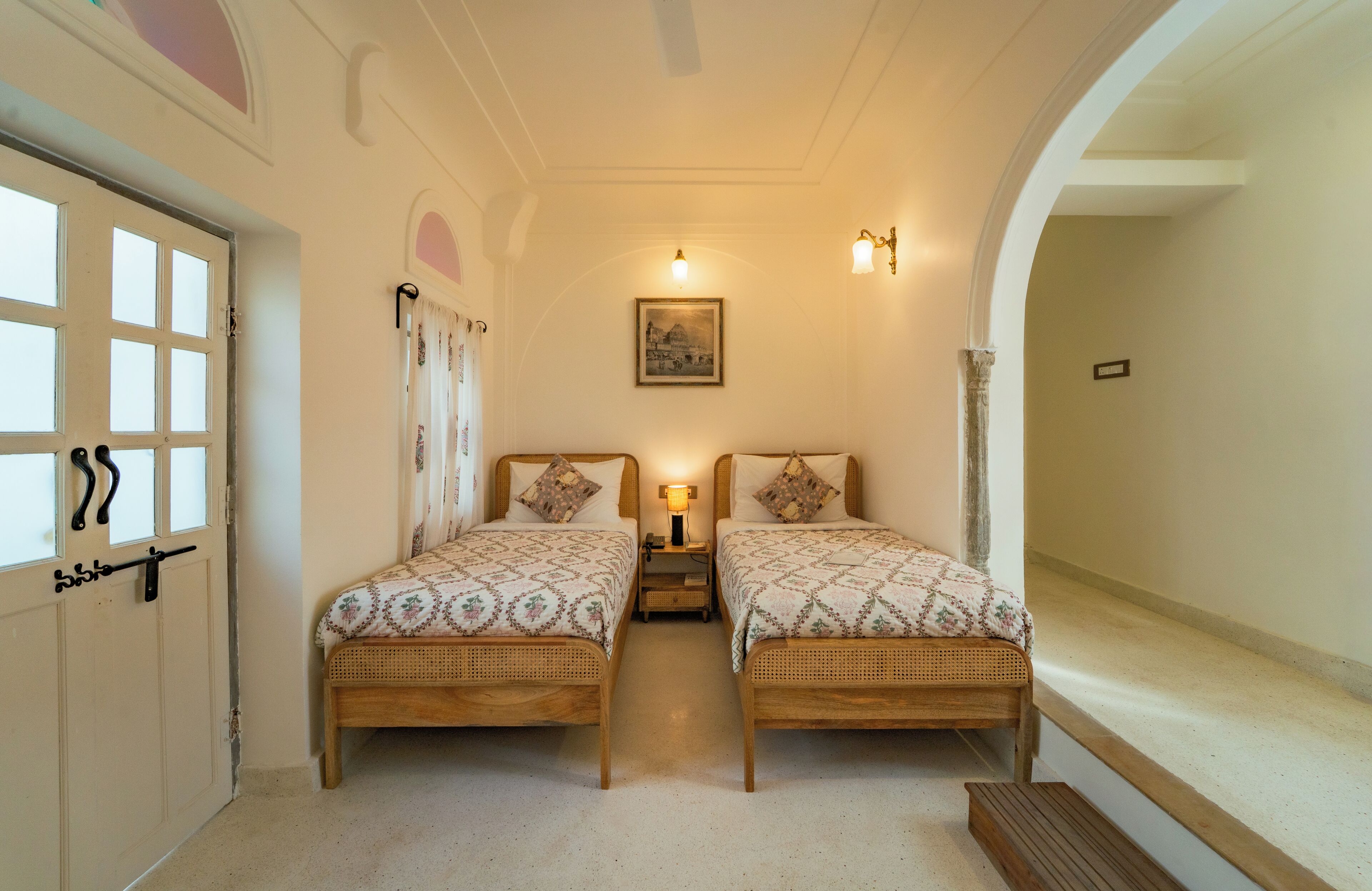 A room with two single beds, a small bedside table with a lamp, a framed picture on the wall, and a door with a handle.