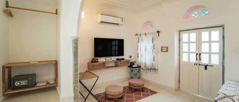 Quarto com televisão de tela plana, prateleira de madeira, mesa pequena, dois pufes, porta e ventilador de teto.