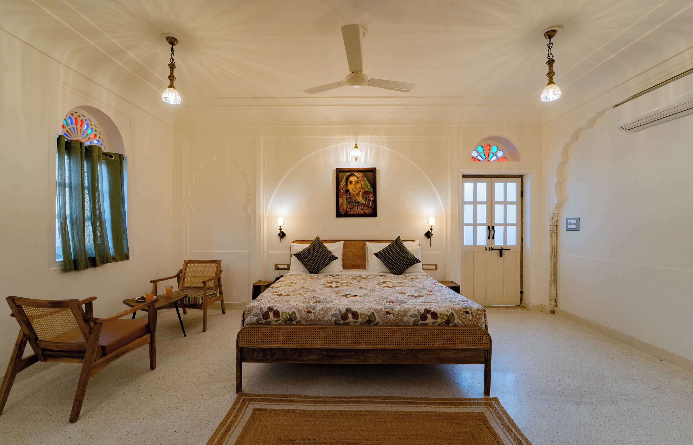 Quarto com cama, duas cadeiras, ventoinha no teto e porta com janela.