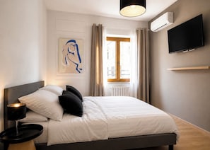 Ein modernes Hotelzimmer mit Bett, Nachttischlampe, einem angebrachten Fernseher und einem Fenster mit Vorhängen.