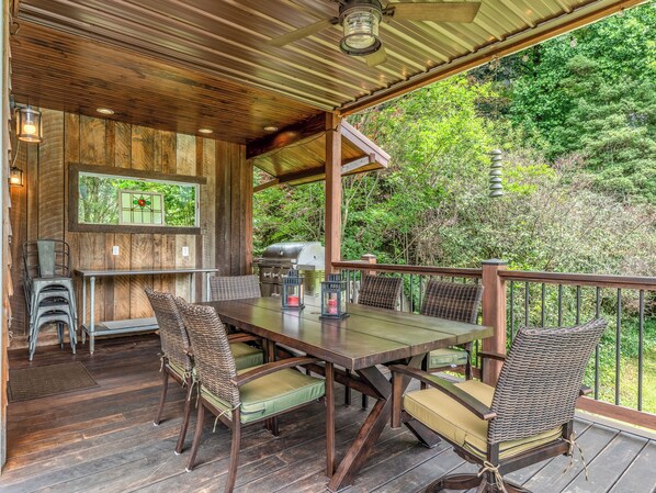 Un área de comedor al aire libre techada con una mesa y sillas de madera, una parrilla y vistas de la vegetación circundante.