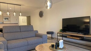 Een moderne woonkamer met een grijze bank, een ronde salontafel, een flatscreen-tv en hanglampen.
