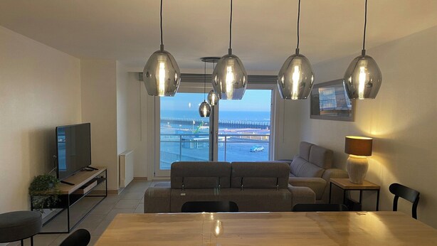 Een moderne woonkamer met een houten eettafel, een bank en een televisie. Er zijn hangende吊灯 en een uitzicht op een brug en water door grote ramen.
