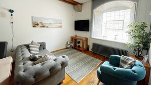 A living room with a grey sofa, a blue armchair, a wooden coffee table, a TV, and a large window with blinds. - Welcome Retreats (Plymouth)