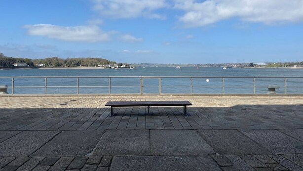 A waterfront promenade with a bench, a railing, and a view of the sea and distant buildings. - Welcome Retreats (Plymouth)