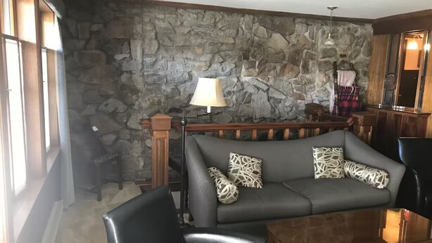 Un salon avec un mur en pierre, un canapé agrémenté de coussins à motifs, un fauteuil en cuir noir et une table basse en bois.