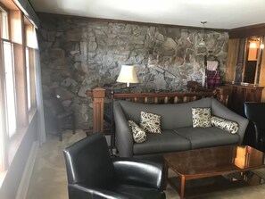 Ein Wohnzimmer mit Steinwand, einer Couch mit gemusterten Kissen, einem schwarzen Ledersessel und einem Holztisch.