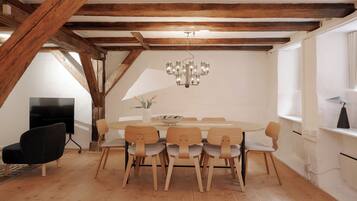 A dining area with a round table, six chairs, a chandelier, wooden beams, a flat-screen TV, and a vase with flowers.