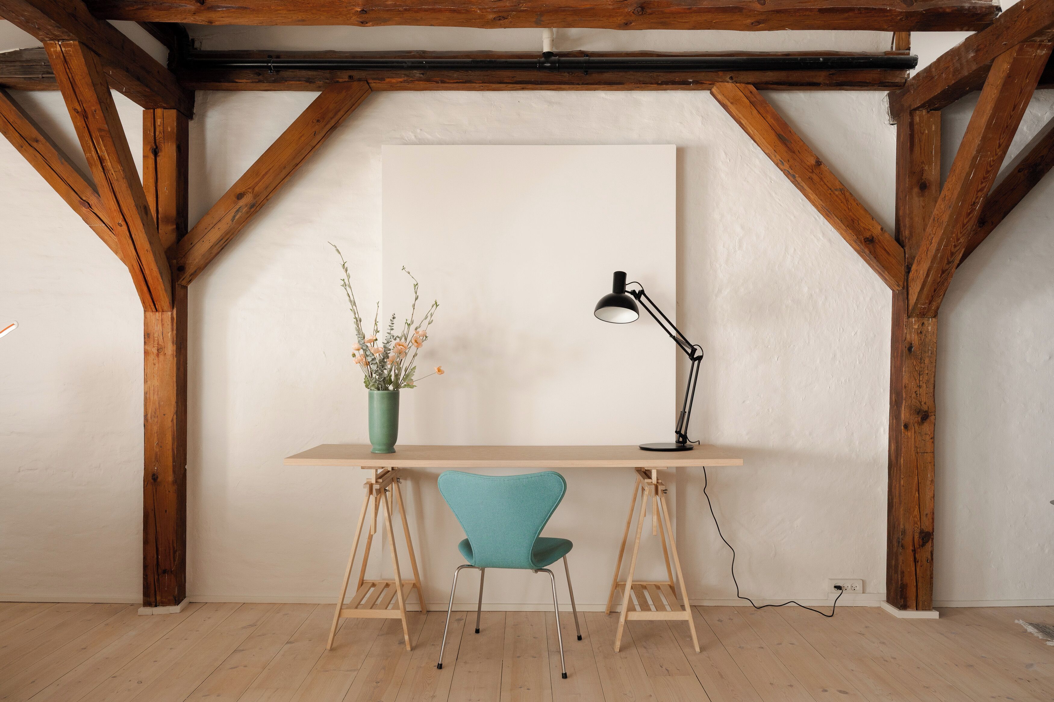 A wooden desk with a teal chair, a black desk lamp, and a vase with flowers.