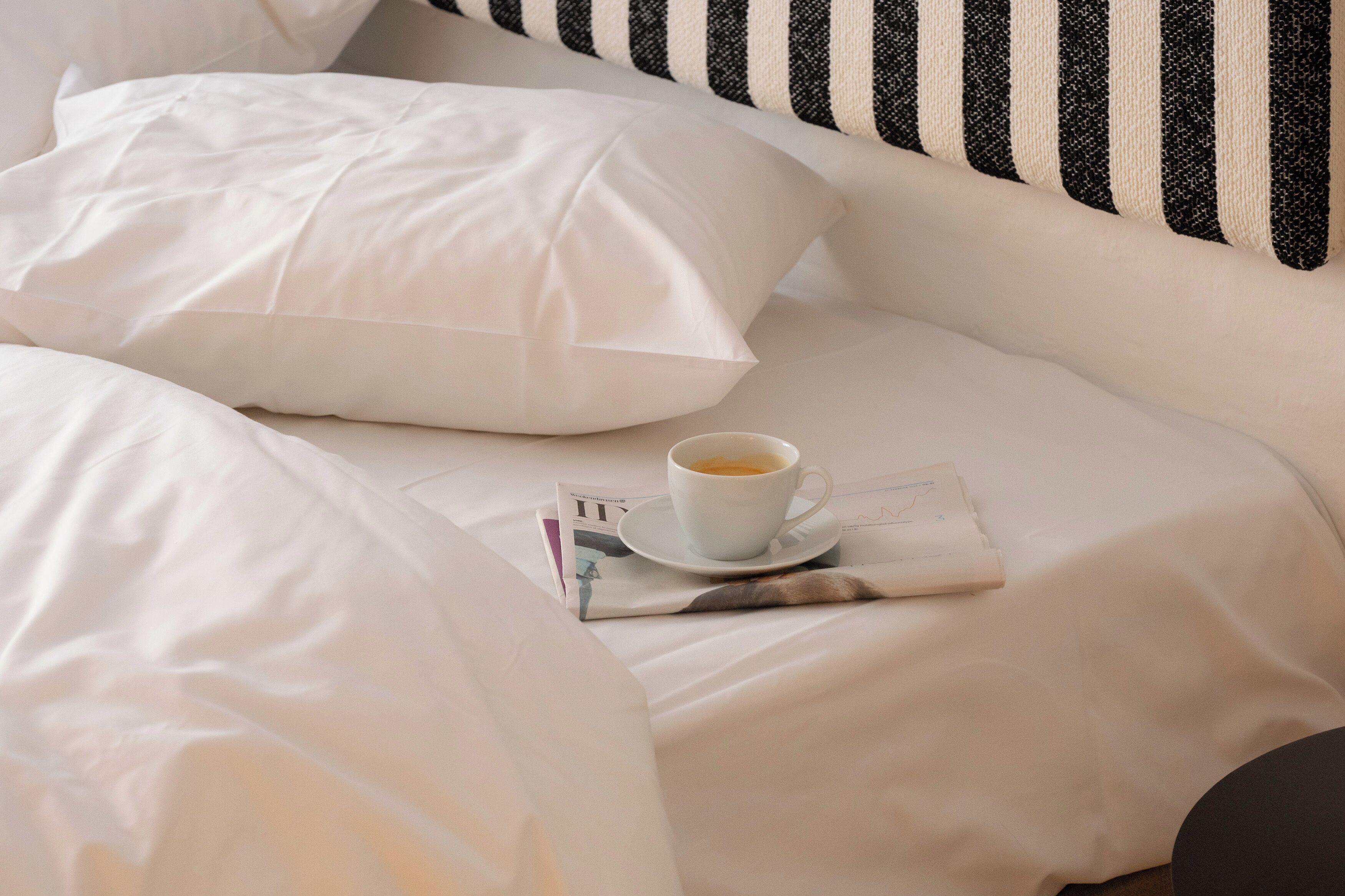 Una cama bien hecha con sábanas blancas, una taza de café sobre un platillo y una revista sobre la cama.