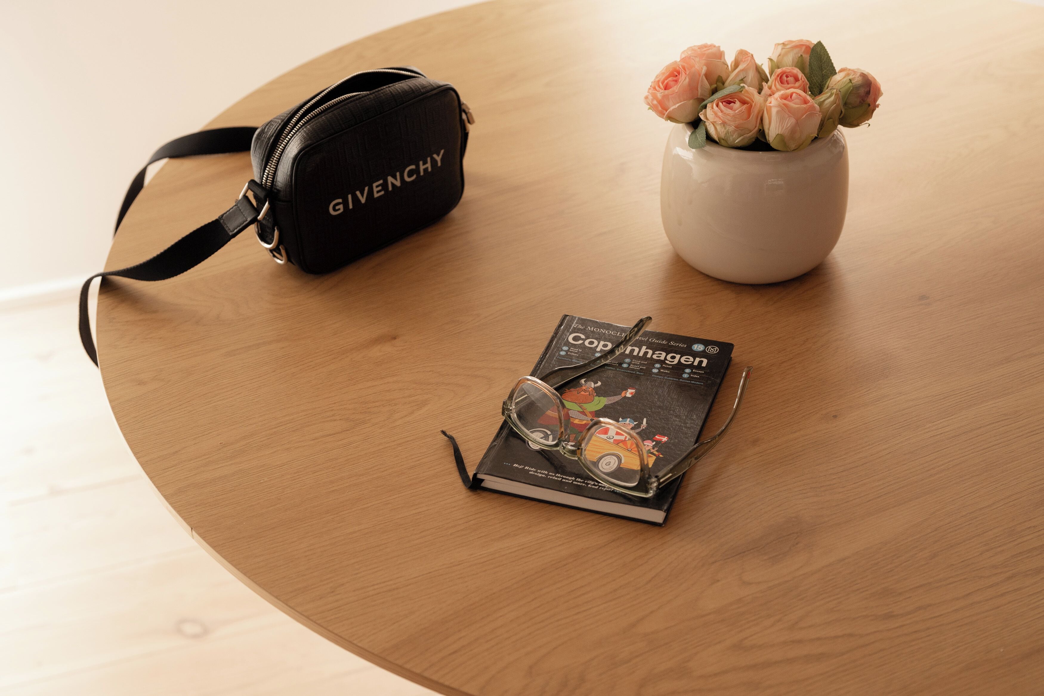 Une table ronde en bois, sur laquelle on aperçoit un sac Givenchy noir, un vase rempli de roses et un livre avec des lunettes posées dessus.