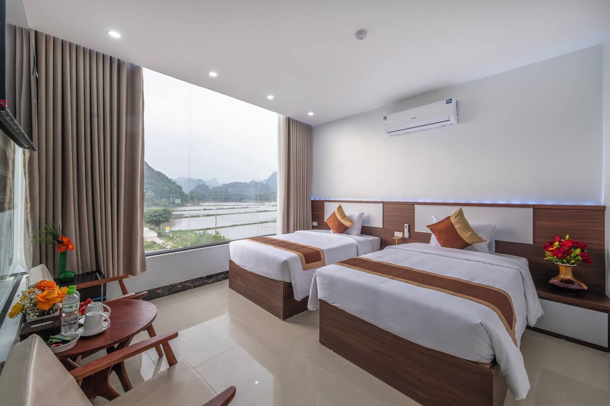 A hotel room with two beds, a dining table, a window with curtains, and an air conditioning unit.