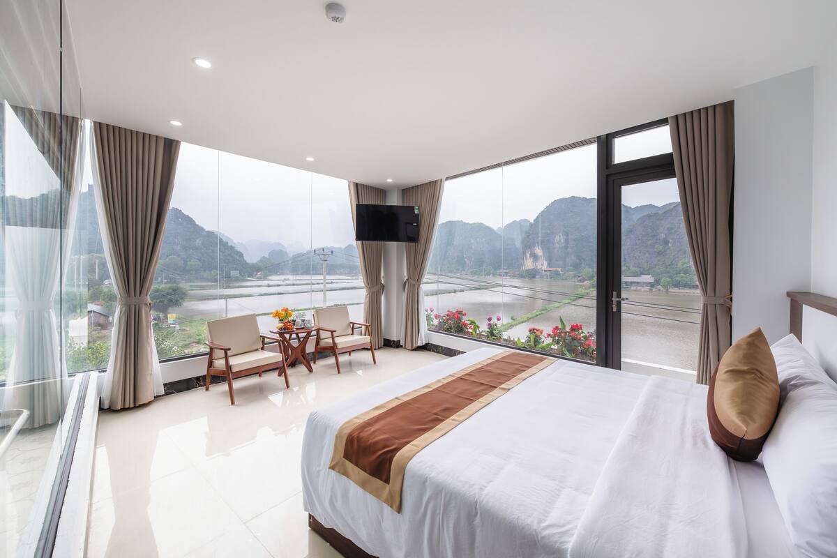 A hotel room with a large bed, a TV, a balcony with a view of mountains and a river, and a seating area with a table and chairs.