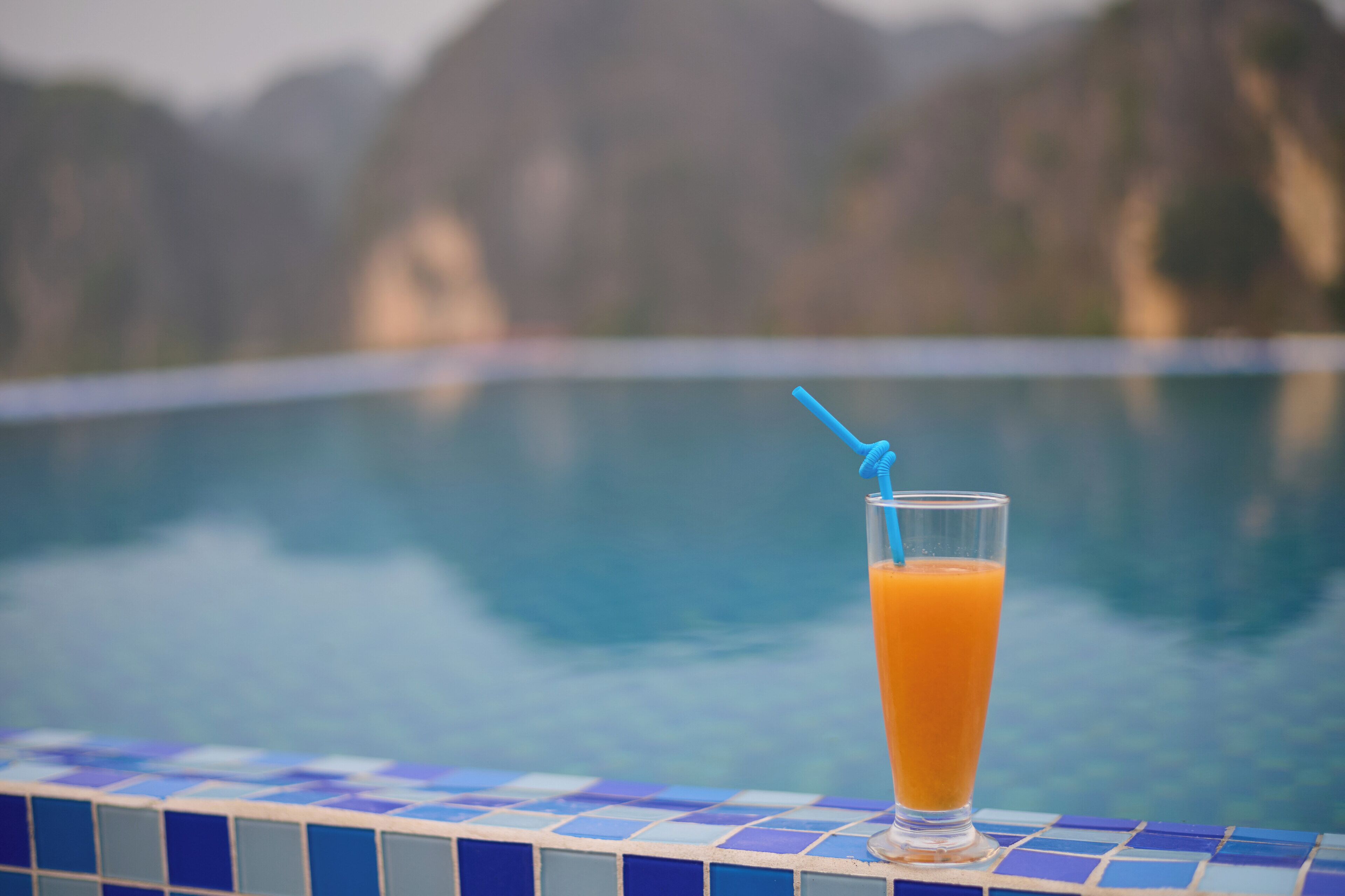A glass of orange juice with a straw on a tiled pool edge.
