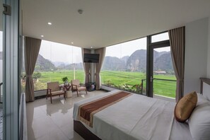 Ein Hotelzimmer mit einem großen Bett, einem Fernseher, einem Balkon mit Blick auf grüne Felder und Berge sowie einer Sitzecke mit Tisch und Stühlen.