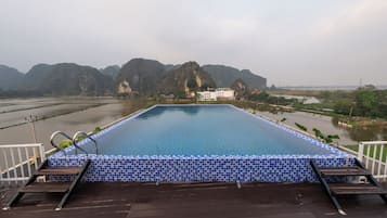 En rektangulär swimmingpool med en blå och vit kakelbeklädd kant, omgiven av ett trädäck med två solstolar och en stege. I bakgrunden syns berg och en sjö eller ett annat vattendrag.