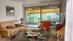 Ein Wohnzimmer mit einer Couch, einem Sessel und einem Couchtisch, einem Ventilator und einem großen Fenster mit Blick auf Grünfläche.