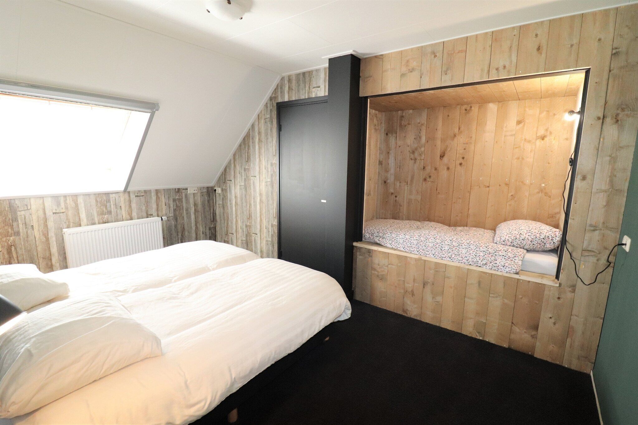 Une chambre avec un plafond en pente, un lit avec une literie blanche, un petit banc en bois avec un coussin et un radiateur.