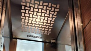 Interior de elevador com paredes metálicas, teto estampado e porta de vidro com um jornal.
