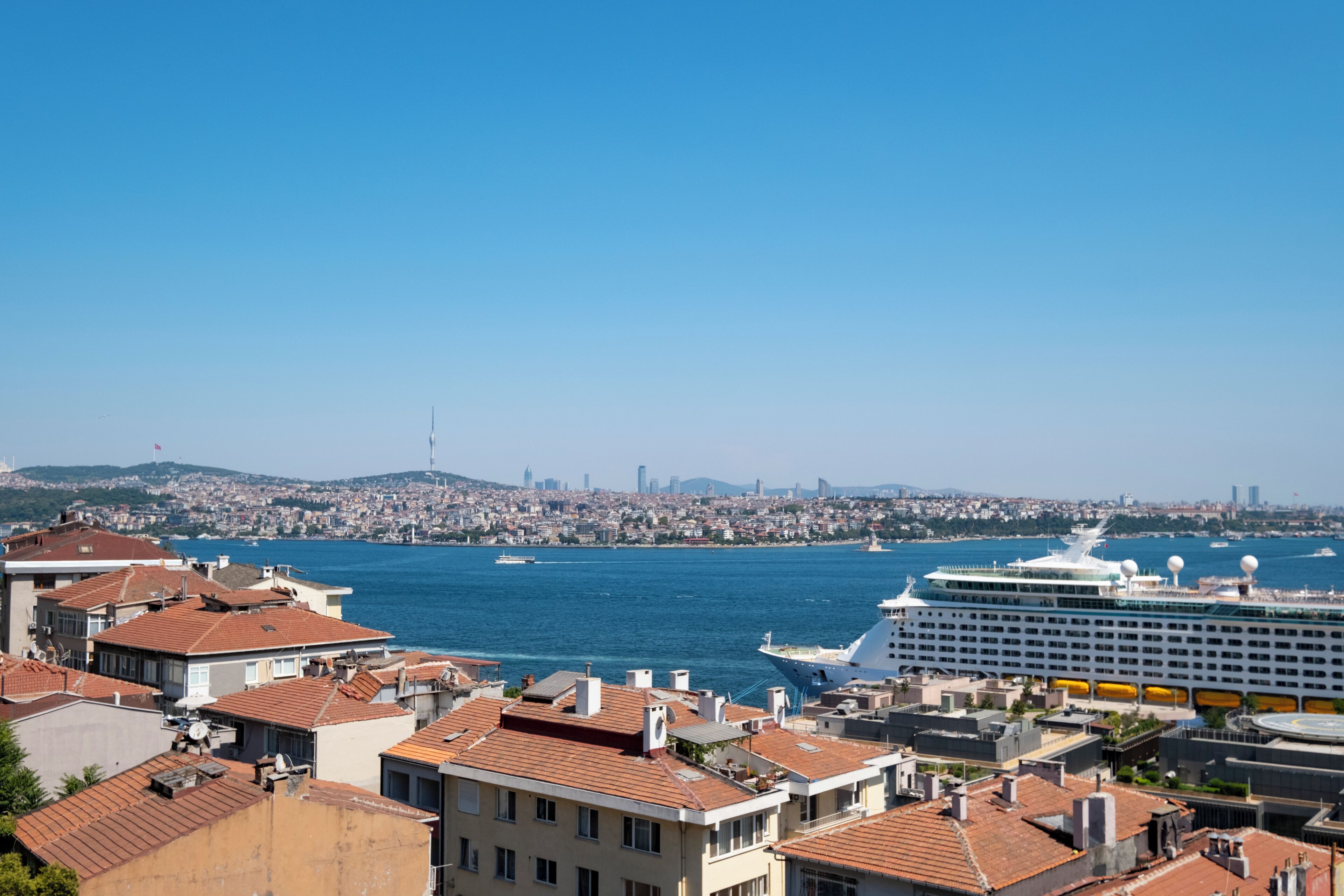 A cruise ship docked near a city with red-roofed buildings and a clear blue sky.