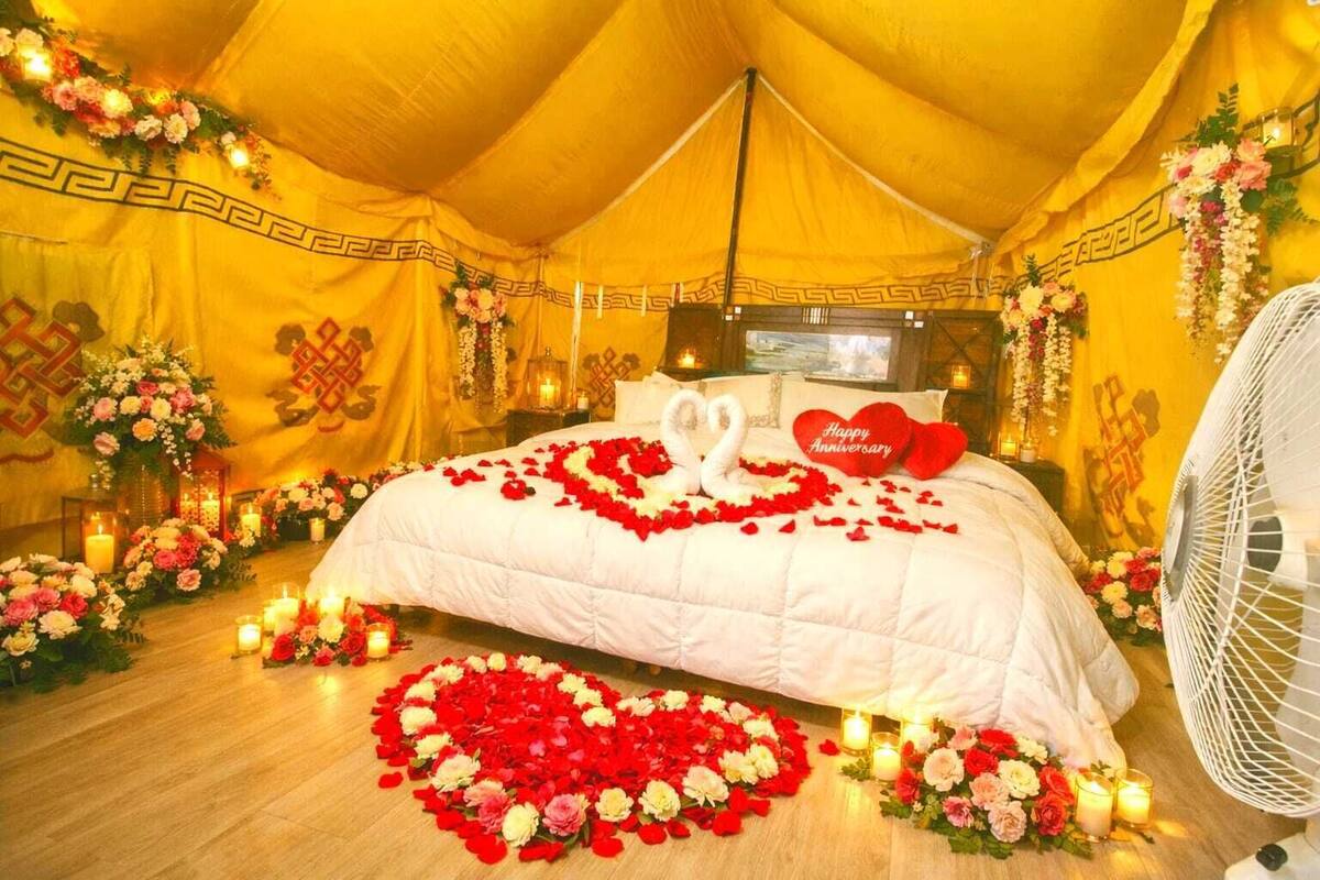 Ein Zeltzimmer mit einem Bett, das mit Rosenblättern und einer herzförmigen Dekoration geschmückt ist, umgeben von Blumenarrangements und Kerzen.