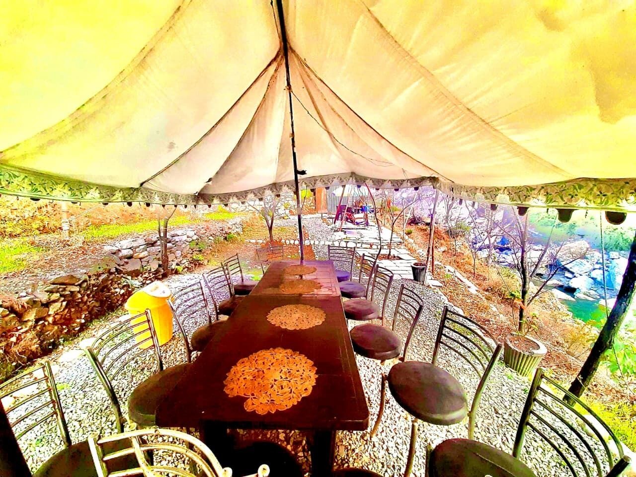 A tented dining area with a long table and chairs, surrounded by a stone wall and a view of a river.
