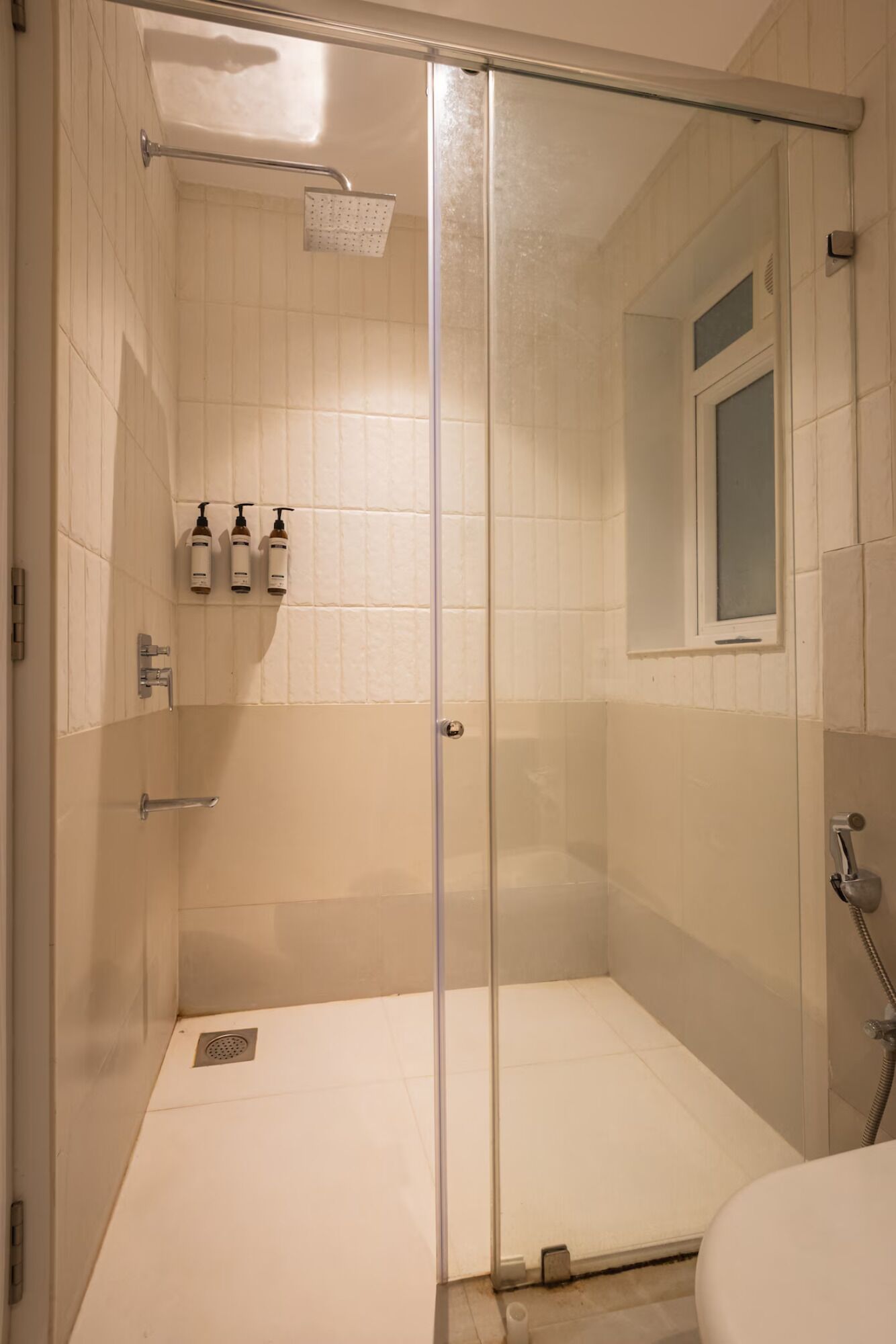 Ducha con mampara de vidrio, pared con azulejos, una ventana y un estante con artículos de aseo.