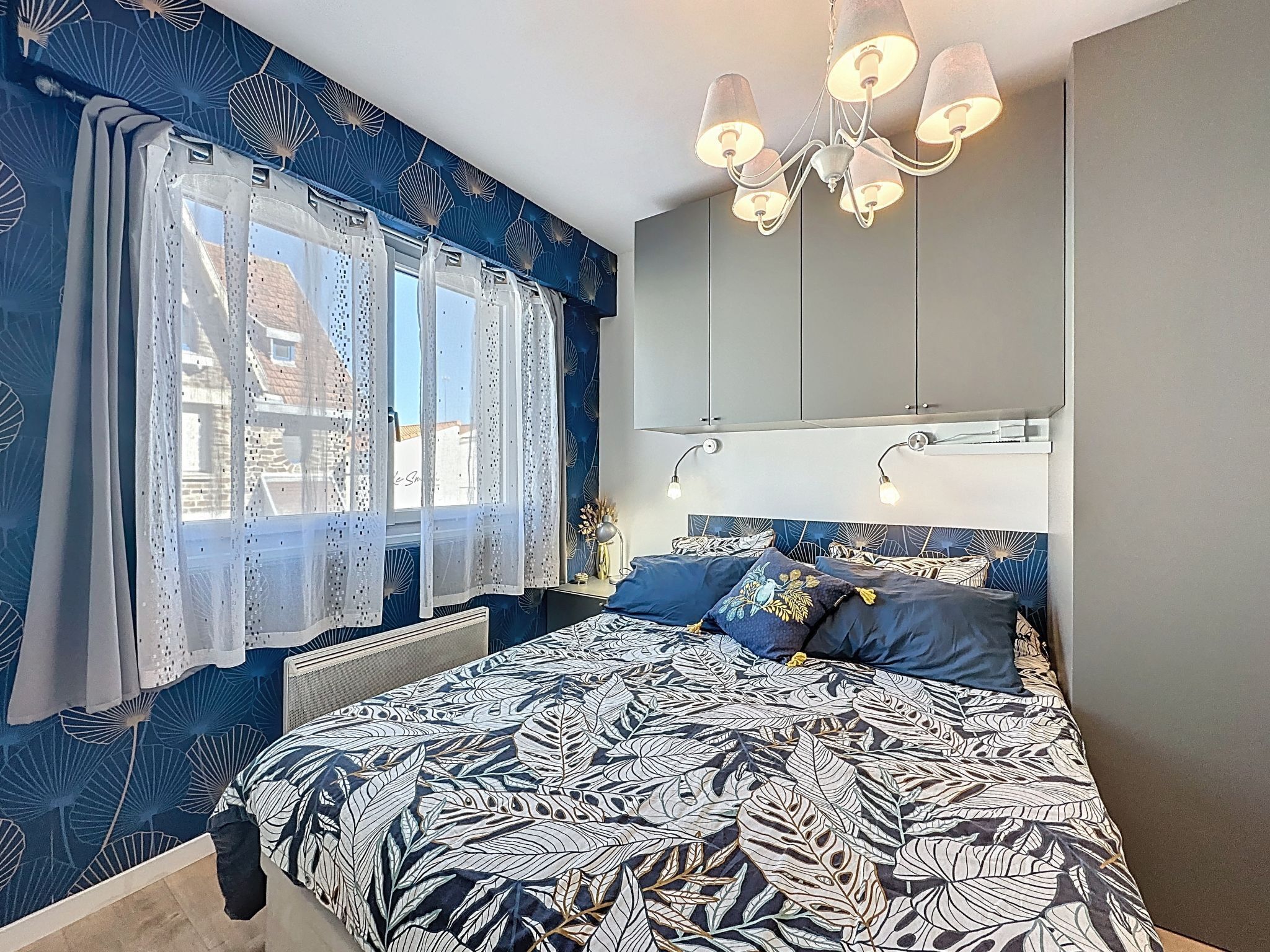 Une chambre à coucher avec un lit, une fenêtre ornée de rideaux légers, un lustre et un mur décoré d’un motif de feuilles.