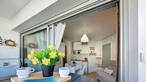 A modern living room with a sofa, a dining table with a magazine, and a potted plant with yellow flowers. - Résidence de la Mer by Interhome (La Bernerie-en Retz)