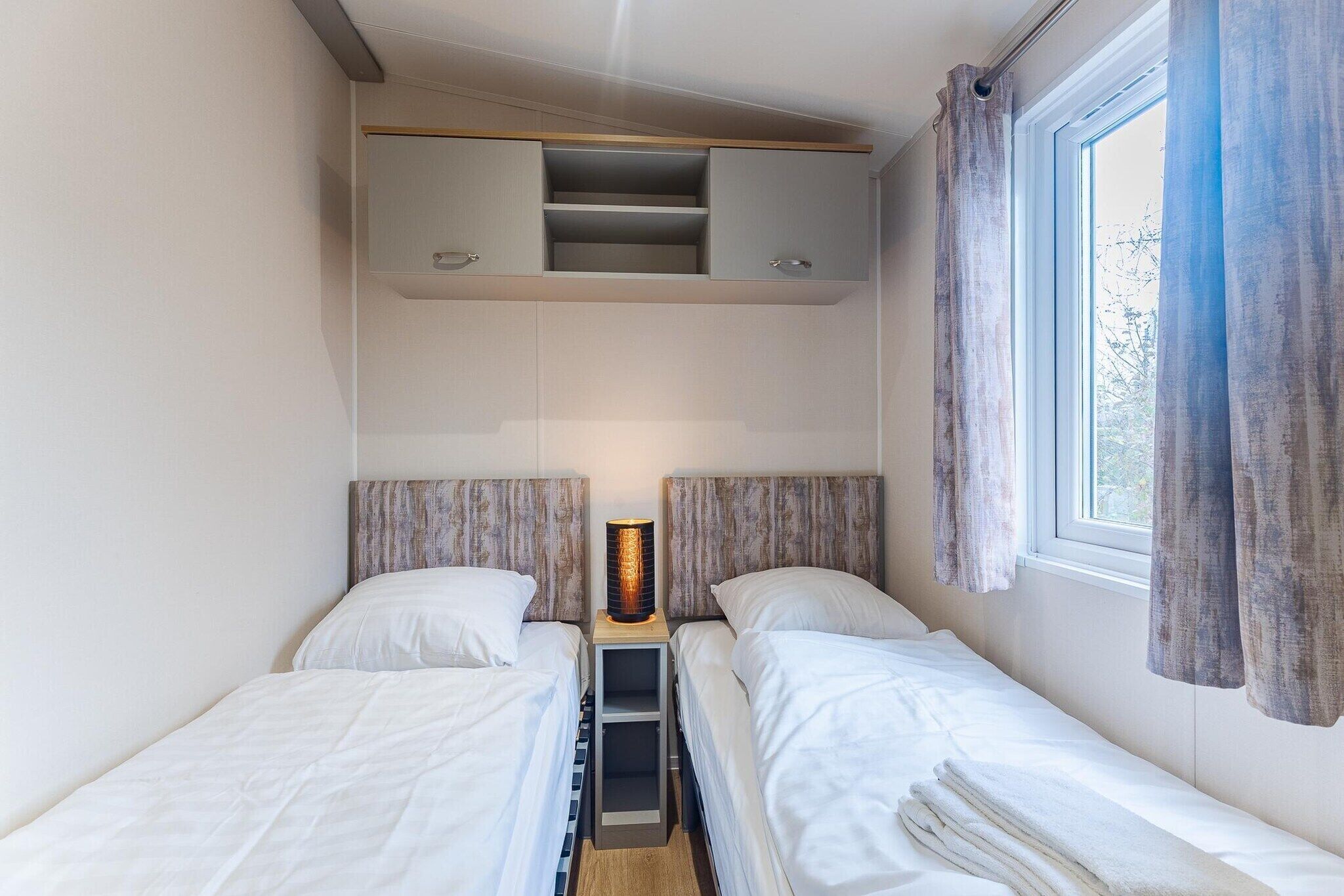 A small, well-lit room with two beds, a bedside table, a shelf unit above the beds, and a window with curtains.