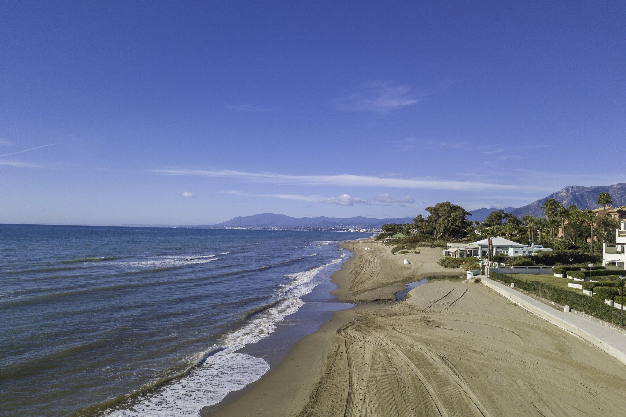 A beach with a clear sky, a sandy shore, and buildings in the background.