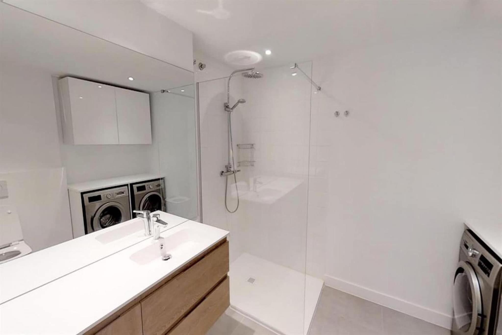 Ett modernt badrum med en dusch som är avskärmad med glasväggar, en vit tvättställ och en inbyggd tvättmaskin.