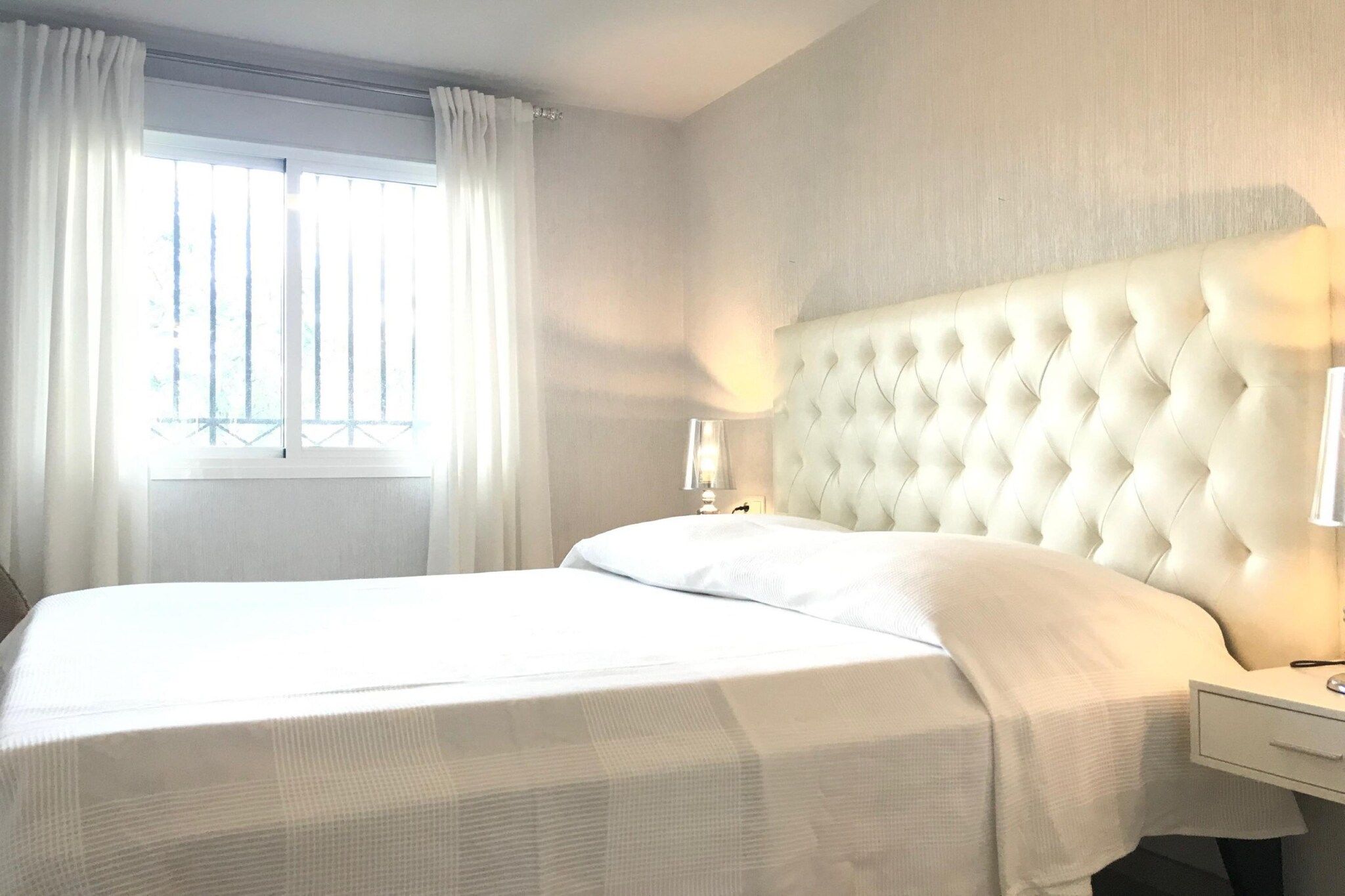 Hotellihuone, jossa on suuri sänky, yöpöytä, lamppu ja ikkuna, jossa on verhot.