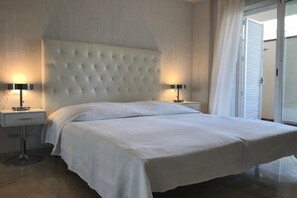 A hotel room with a large bed, two bedside tables with lamps, a tufted headboard, and a view of the outside through a window with blinds. - Cozy Elviria Beachside Apartment (Marbella)