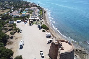 Vue aérienne d’une zone côtière avec une plage, des véhicules garés et des bâtiments.