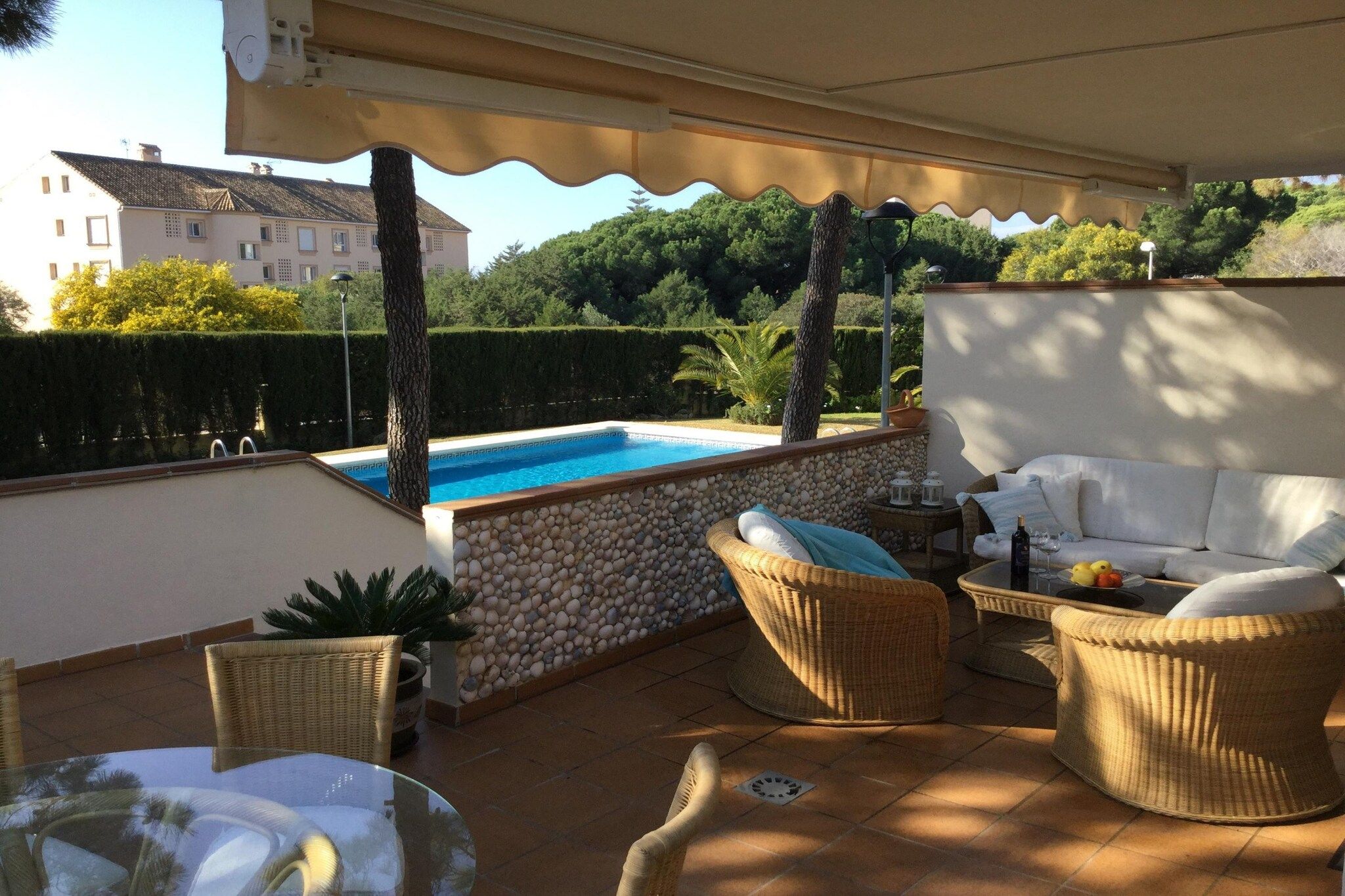 A covered outdoor area with a pool, wicker furniture, and a view of a building in the background.