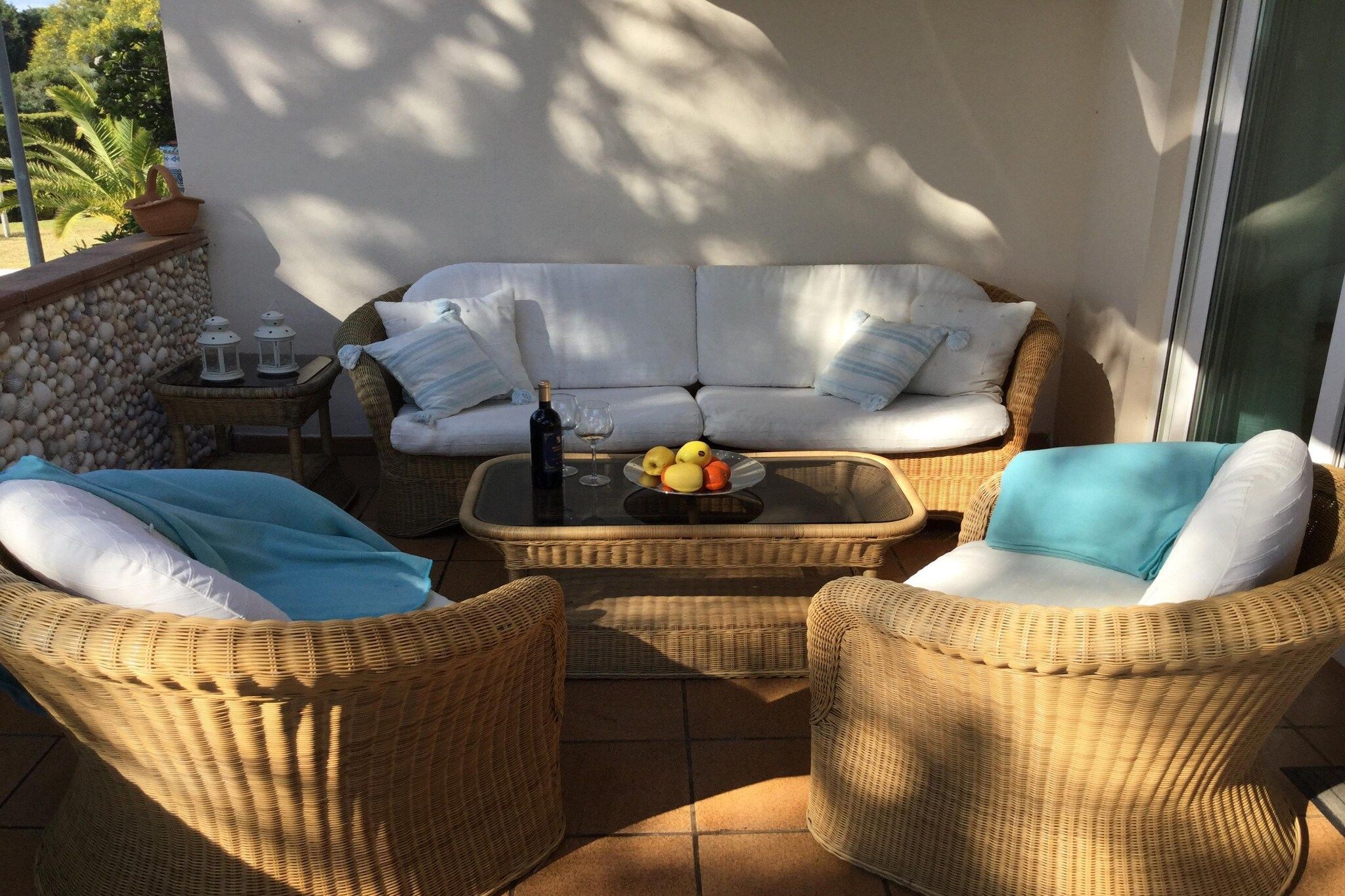Un conjunto de sofá y sillones de mimbre con cojines, una mesita con un frutero y una botella de vino.