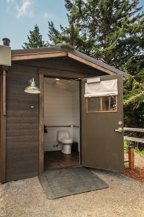 Una pequeña cabaña rústica de madera, con un inodoro y una ventana con cortina blanca.