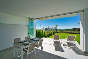 A modern dining area with a table set for four, overlooking a lush green landscape and a clear blue sky. - Crystal Lagoon Apartment Retreat (Casares)