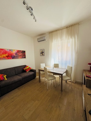 A living room with a sofa, a dining table with chairs, an air conditioning unit, and a window with curtains. - Your vacation home between technology and the sea, just a few steps from Ostia beach (Lido di Ostia)