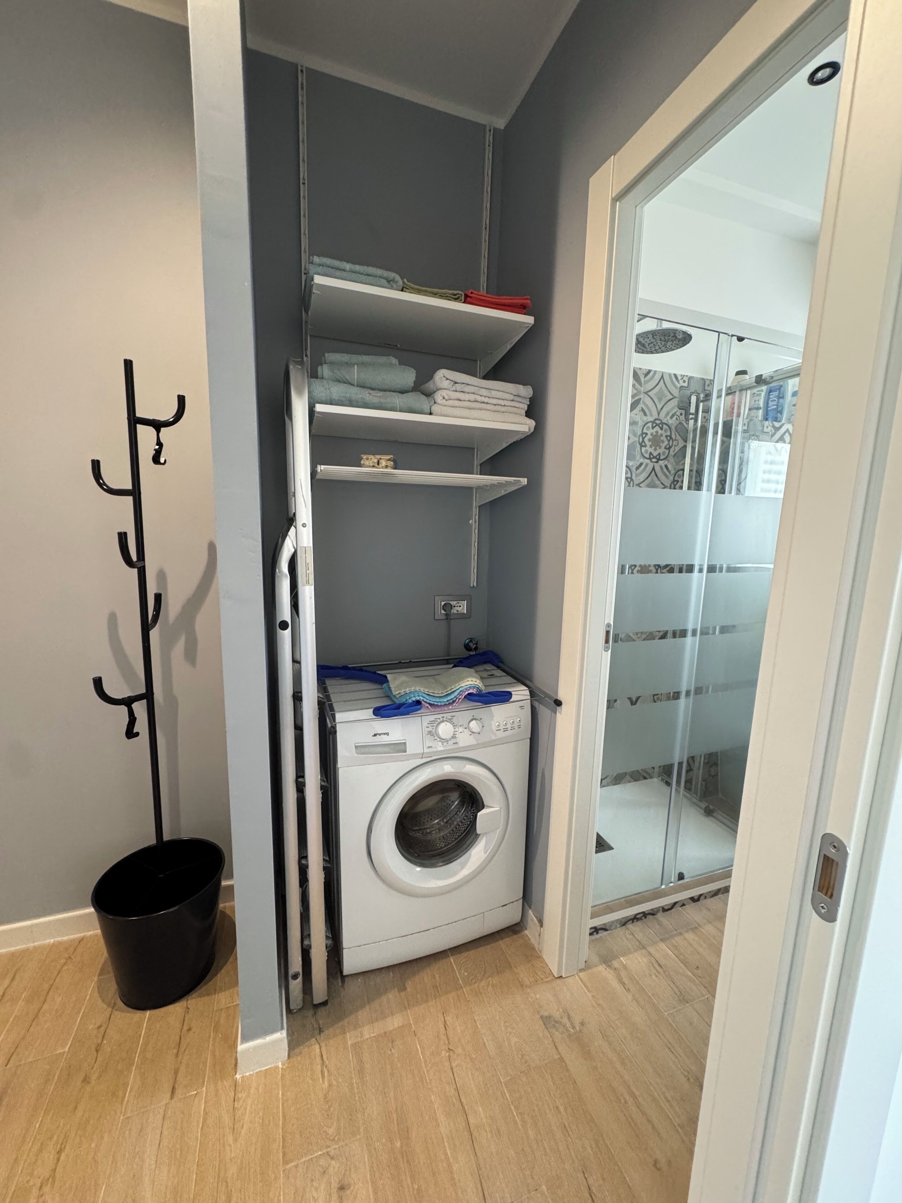 A laundry room with a washing machine, shelves with folded towels, and a coat rack.