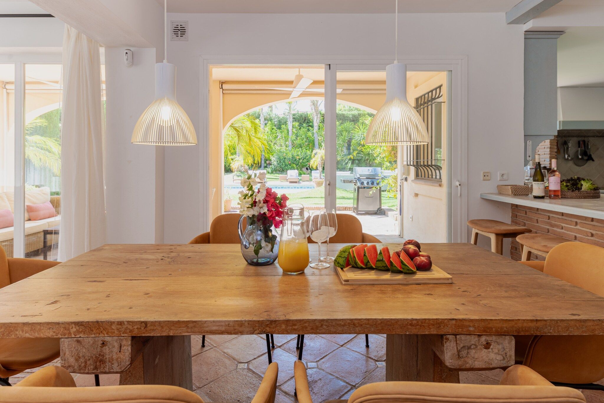 A dining area with a wooden table, chairs, a vase with flowers, a fruit platter, and a view of a garden through a glass door.