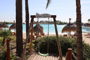 Een strandgedeelte met een schommel, palmbomen en een bord waarop staat 'Alcazaba Vive Lagoon'.