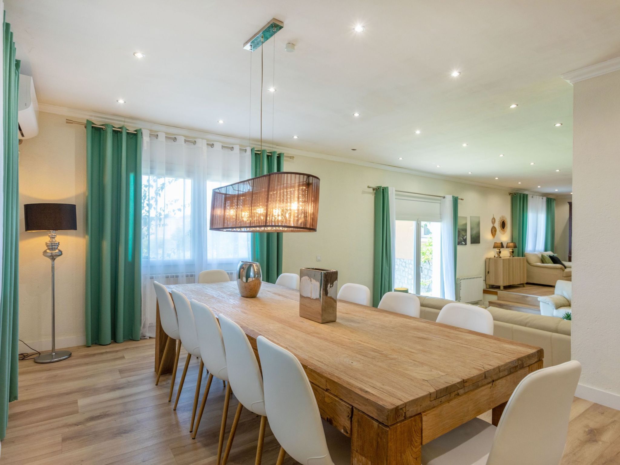 Een eetgedeelte met een houten tafel, witte stoelen, een hangende lamp en uitzicht op een woonkamer met een bank en salontafel.