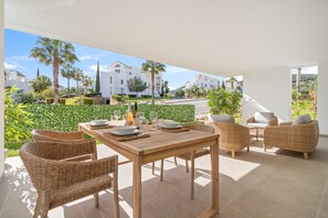 Une terrasse avec une table en bois dressée pour le repas, des chaises en osier et une vue sur un quartier résidentiel agrémenté de palmiers et de bâtiments blancs.