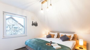 Ein Schlafzimmer mit einem großen Bett, einem Nachttisch mit Lampe, einem Fenster mit Blick auf ein Haus und einer Wandleuchte.