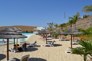 A beach with sun loungers and thatched umbrellas, a pool, and a building in the background. - Elegant 4-Pax Apartment in Casares (Casares)