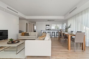 A modern living room with a sofa, coffee table, and TV. There is a dining area with a table and chairs. The room has white walls and a wooden floor. - Spacious Apartment for 4 with Solarium (Casares)